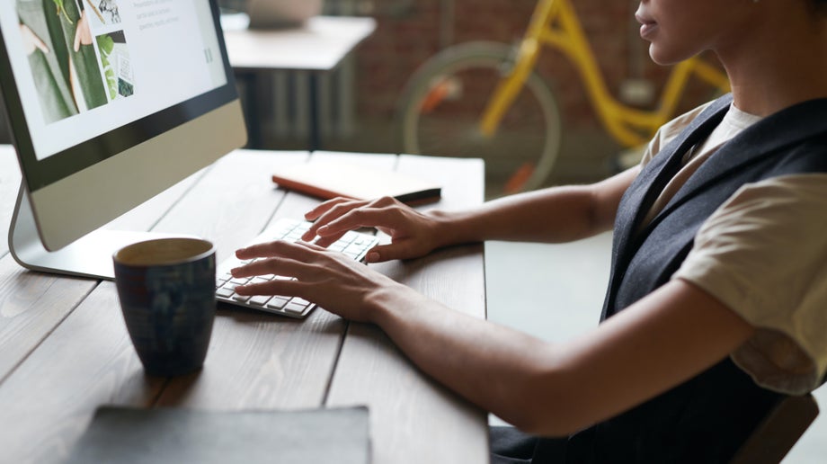 A designer is working at their desk. Their hands are on the keyboard and visuals are displayed on the computer screen.