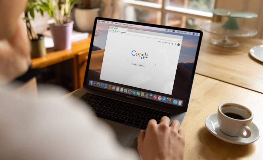 The Google homepage appears on someone's laptop as they drink a coffee at a table 