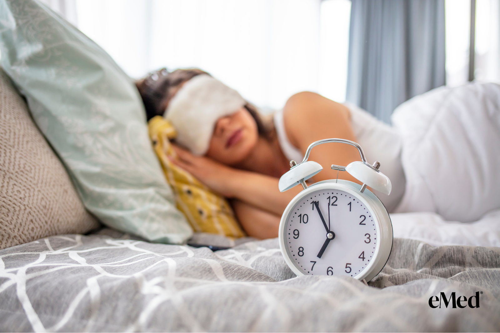 A woman sleeping on a bed with her eyes covered by a sleep mask, and in front of her on the bed is an old-fashioned alarm clock