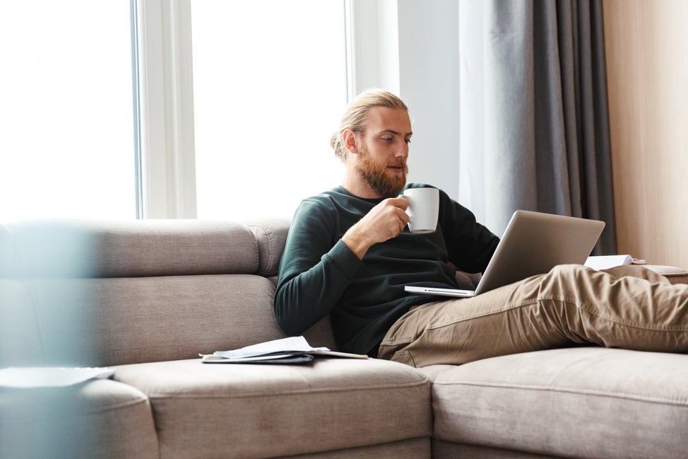 Man drinks coffee on couch while looking at computer