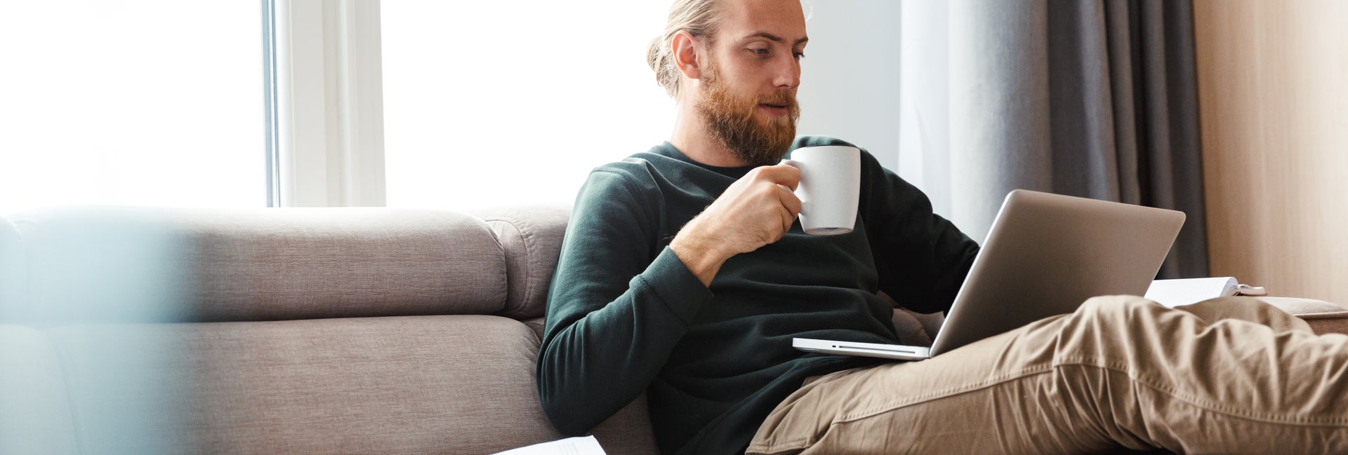 Man drinks coffee on couch while looking at computer