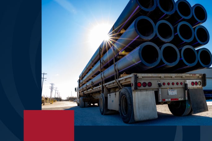 A flatbed truck loaded with large black polyethylene pipes, stacked horizontally, parked on a sunny day. The sunlight creates a flare behind the pipes, and the blue sky contrasts with the industrial setting. A red and blue graphic element is visible in the corner.