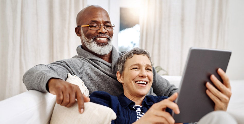A couple sitting on a couch smiling while using a tablet. 