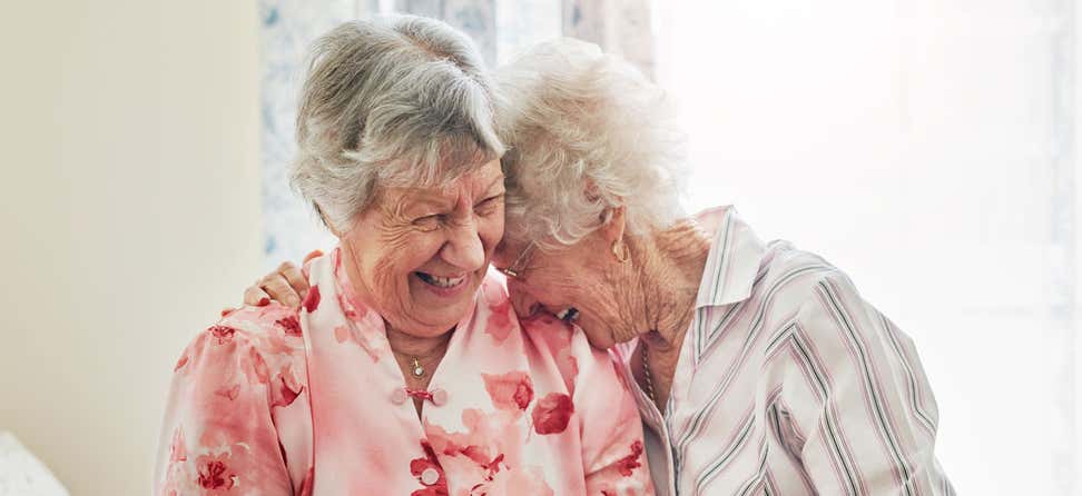 Two Caucasian senior female friends are laughing and hugging, sharing a joke together.