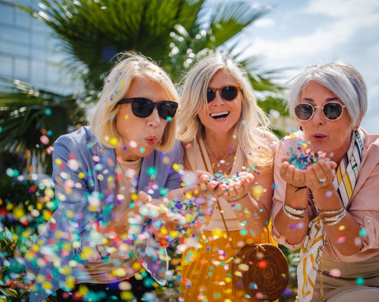 Three senior women wearing sunglasses are outside in the sun, blowing confetti while at a birthday party.