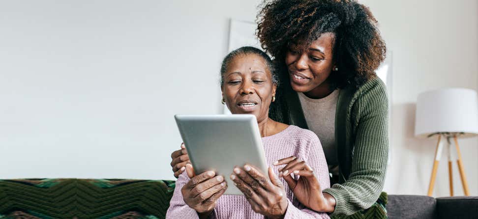 A younger caregiver is helping her older adult mom use her tablet while sitting on the couch.