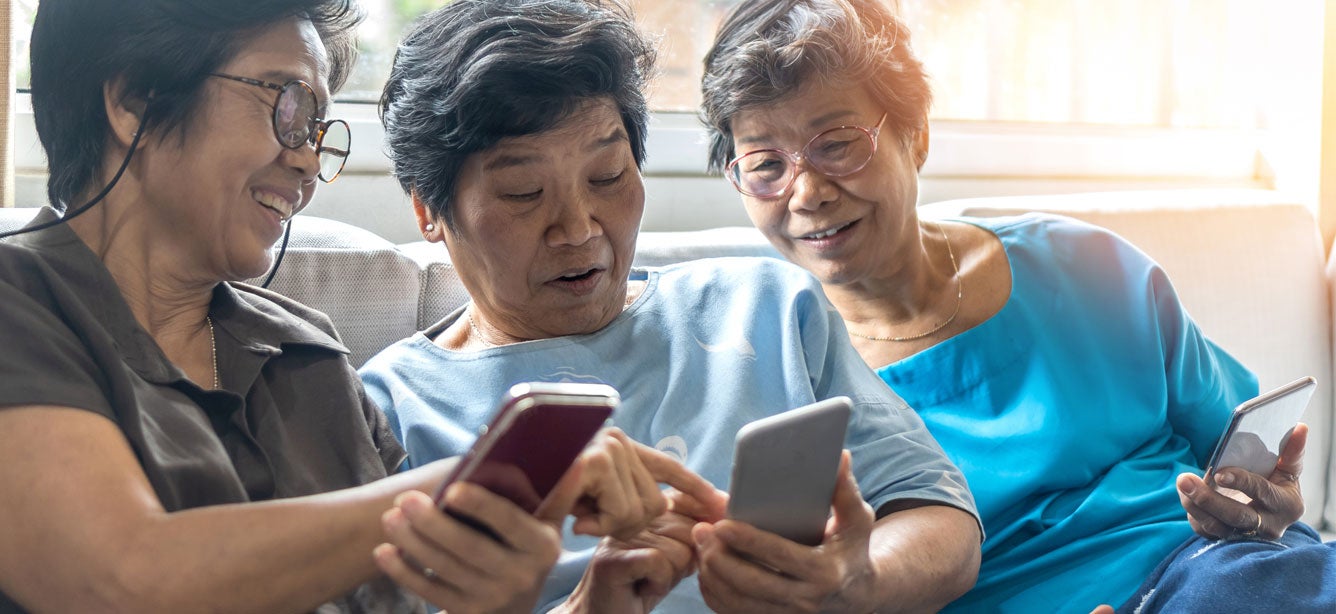 A group of senior Asian women are comparing cell phones while sitting together on a couch.