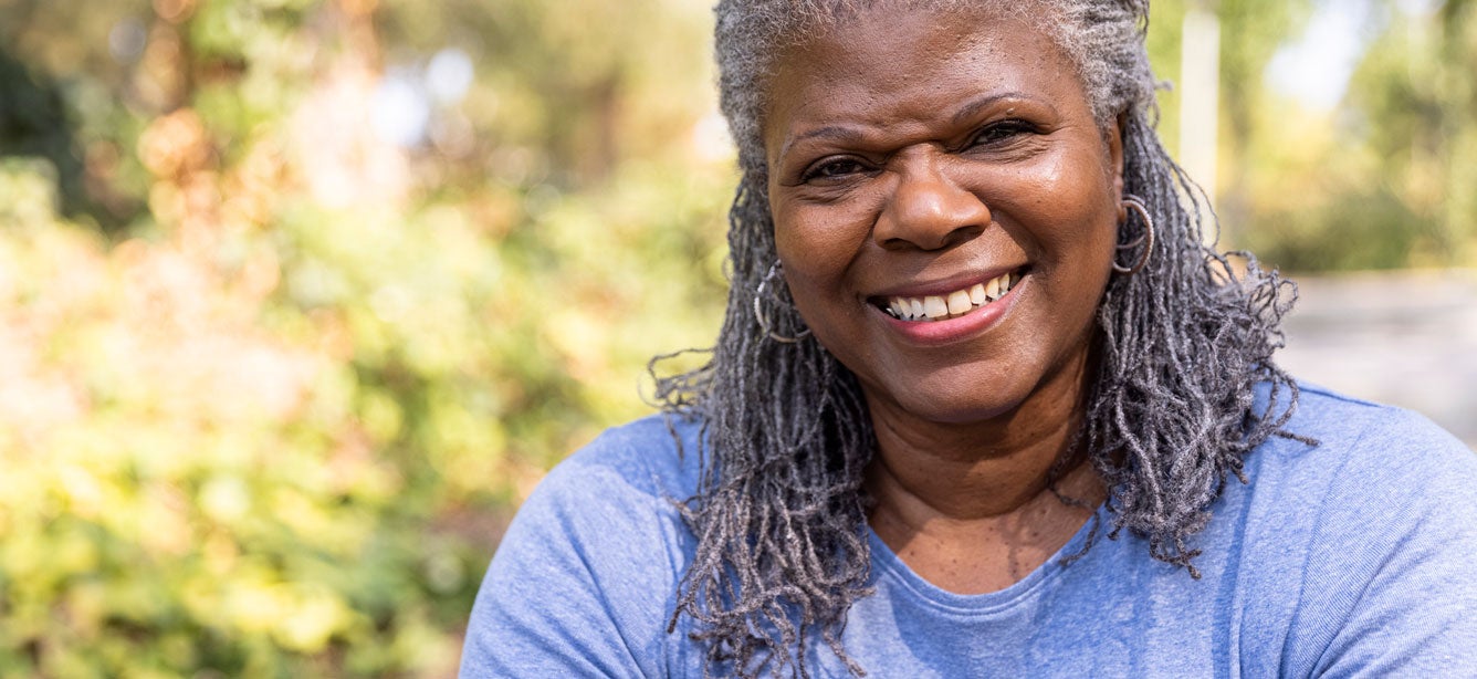 An up close shot of a large senior Black woman wearing a blue t-shirt, sitting outside smiling for the camera.