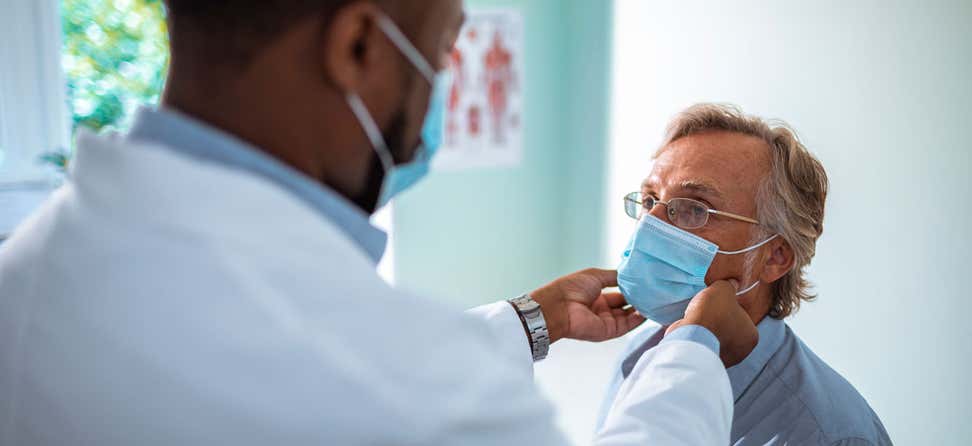 A senior Caucasian man is at his doctor's office receiving a wellness exam. The Black doctor is examining the man's throat.