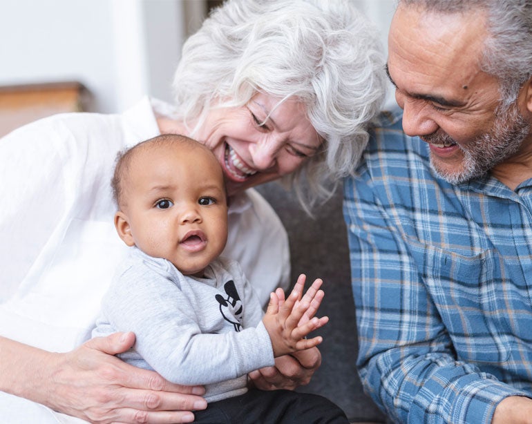 A mixed-race senior couple is holding their grandchild and laughing together.