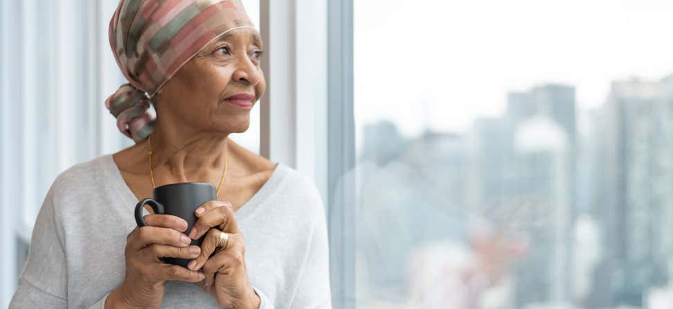 A black senior woman with cancer is wearing a scarf on her head, staring out the window of her hospital room.