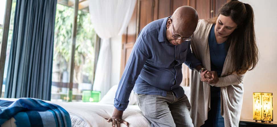 A homecare worker is helping a senior Black man get up and out of bed.