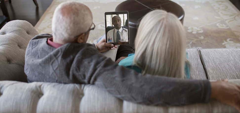 Older couple sitting on couch talking with doctor via telemedicine appointment on tablet