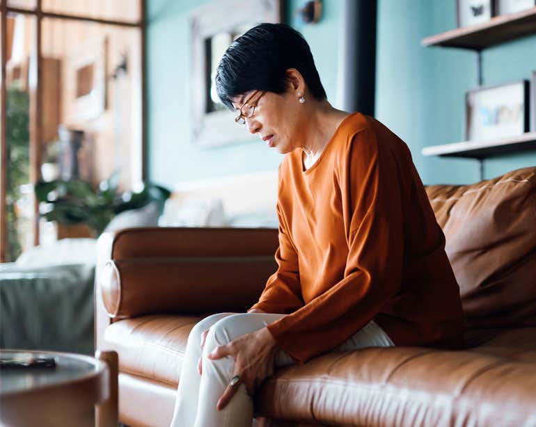 A senior Asian woman is holding her leg in pain as she sits on the couch. Diminished bone density can lead to osteoporosis—a debilitating disease that increases the risk of fractures. Get the information to stay proactive about your bone health.