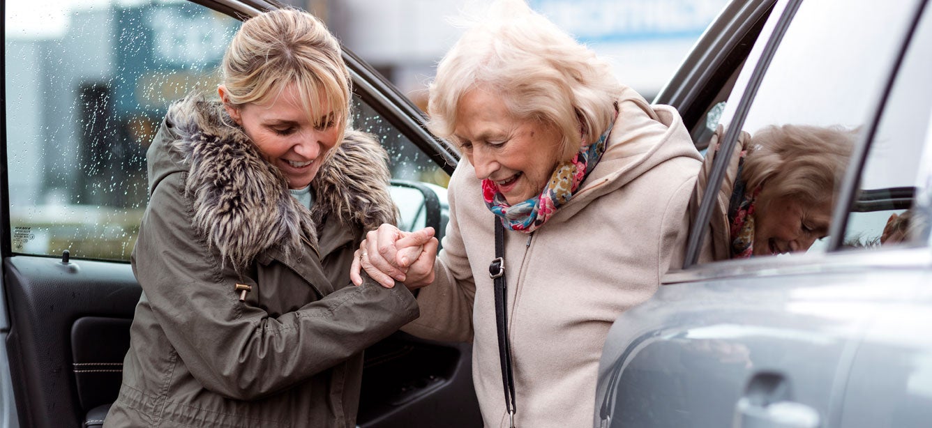 A young female caregiver helps a senior woman out of the passenger seat of her car.