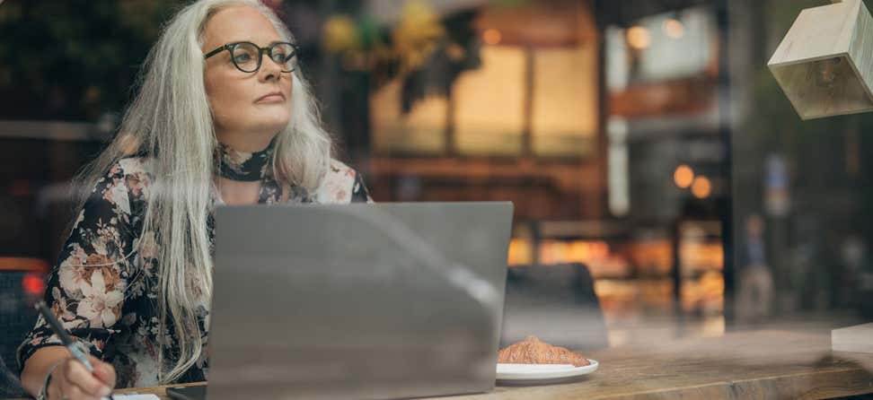 A senior woman wearing glasses sits at her computer in a coffee shop, contemplating a decision and looking out the window.