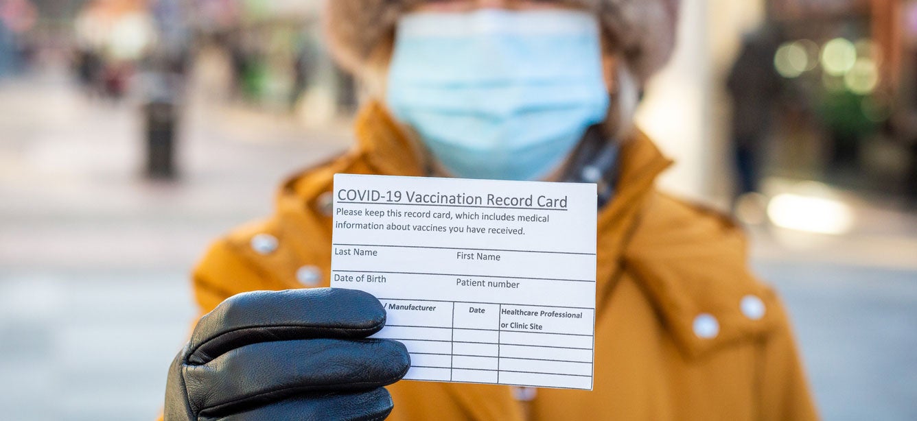 A senior woman dressed in a winter coat, hat, and gloves is holding her COVID-19 vaccination card out for a close up shot. 