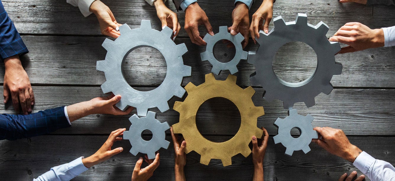 Several hands are holding large cogs in the center of a table, indicating teamwork and having to work together.