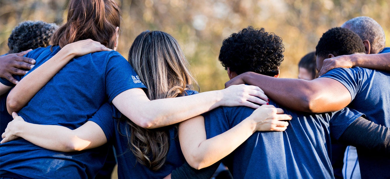 A diverse group of people from the back are in a huddle with their arms around each other's shoulders.