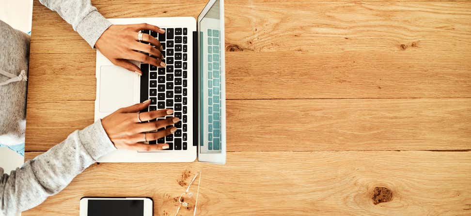 A Black woman, pictured from above, is writing a blogpost on her laptop computer while listening to music.