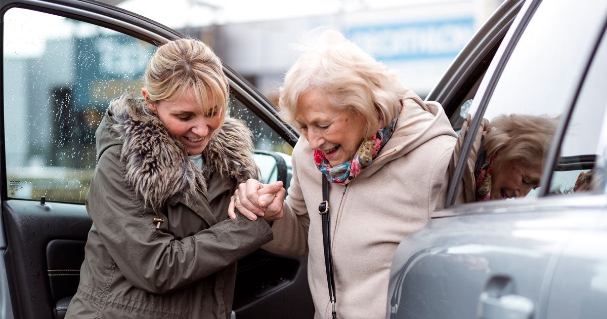 A young female caregiver helps a senior woman out of the passenger seat of her car.