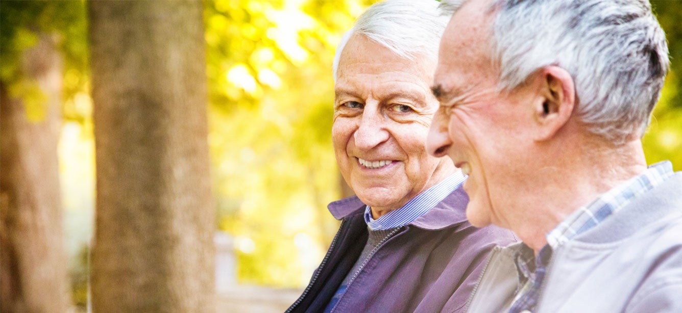 A senior Caucasian gay couple are laughing together as they take a stroll through the park.