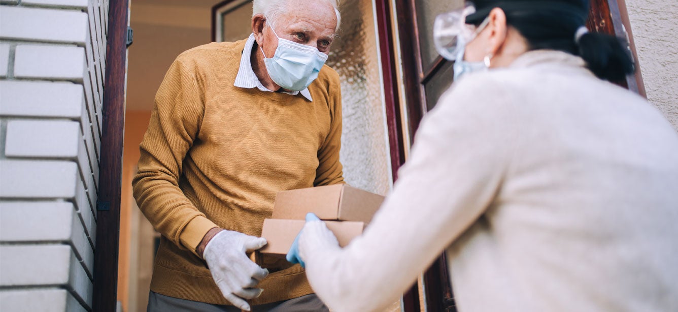 A food delivery service drops of groceries to a senior man wearing a mask and gloves during the pandemic.