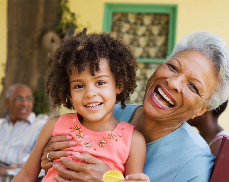 A senior Black woman is holding her granddaughter in her lap during a family get together outside.