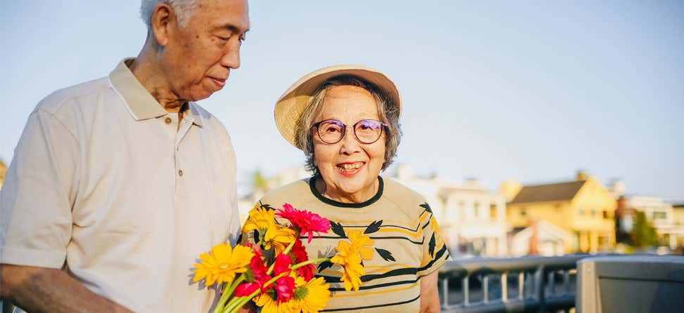 A senior Asian couple walks hand-in-hand outside, where the husband is carrying a bouquet of flowers as the wife smiles at the camera.