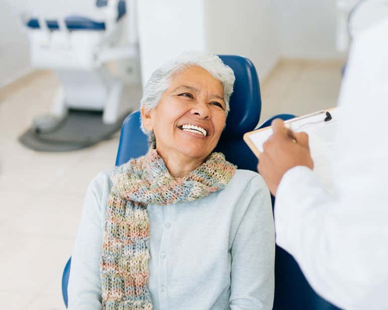 A Hispanic older woman is sitting in a dental office chair, talking to her dentist.