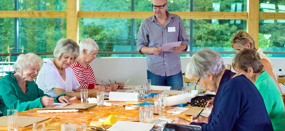 A group of seniors are painting in an art class with their middle-aged instructor who's wearing glasses.