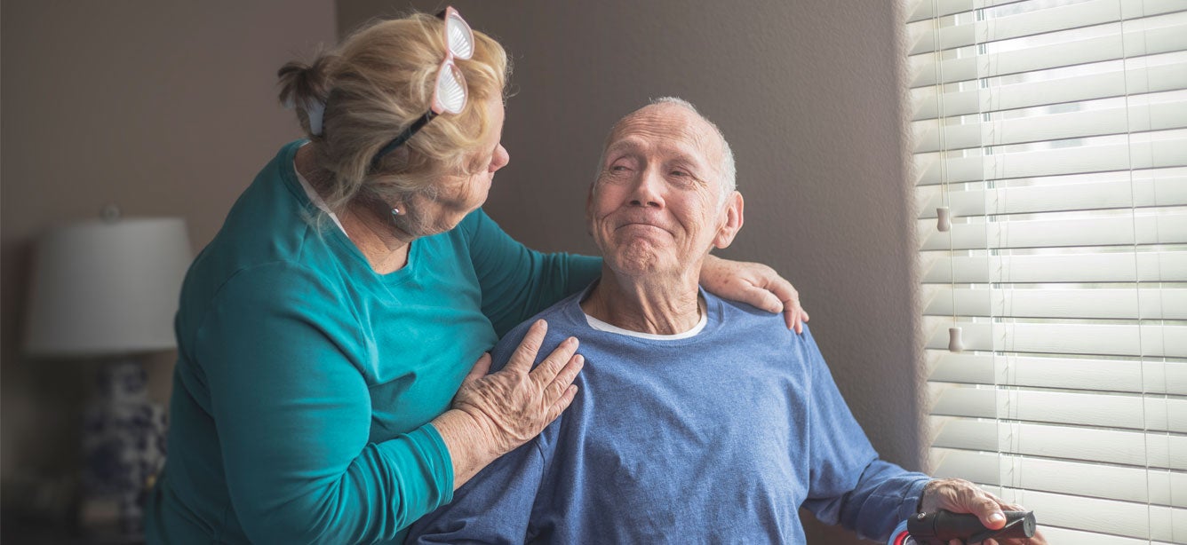 An older female caregiver looks fondly on a much older senior man who's sitting down, smiling by a window.