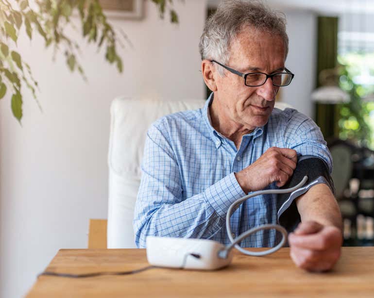 A senior man is sitting down at the kitchen table using a medical device to measure his blood pressure.