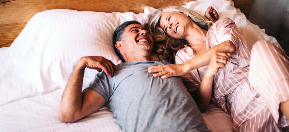 A senior Caucasian couple lays in bed in their pajamas, laughing together.