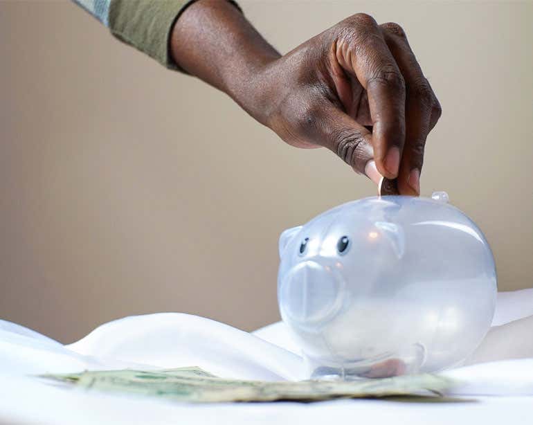 A Black senior hand is placing coins in a clear piggy bank.
