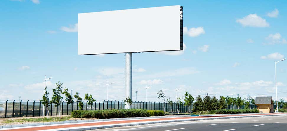 A blank billboard near a highly trafficked road stands blank against a blue sky.