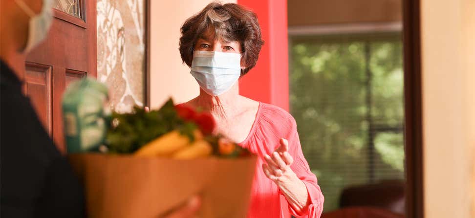 A senior masked woman stands at the door of her house receiving a grocery delivery.