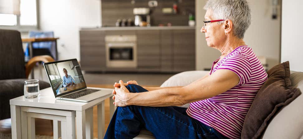 A senior woman is sitting on the sofa in her living room having a video call with her health advisor.