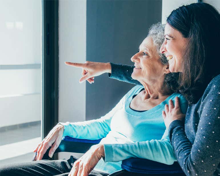 An older female caregiver has her arms around her senior female loved one as they both stare happily outside.