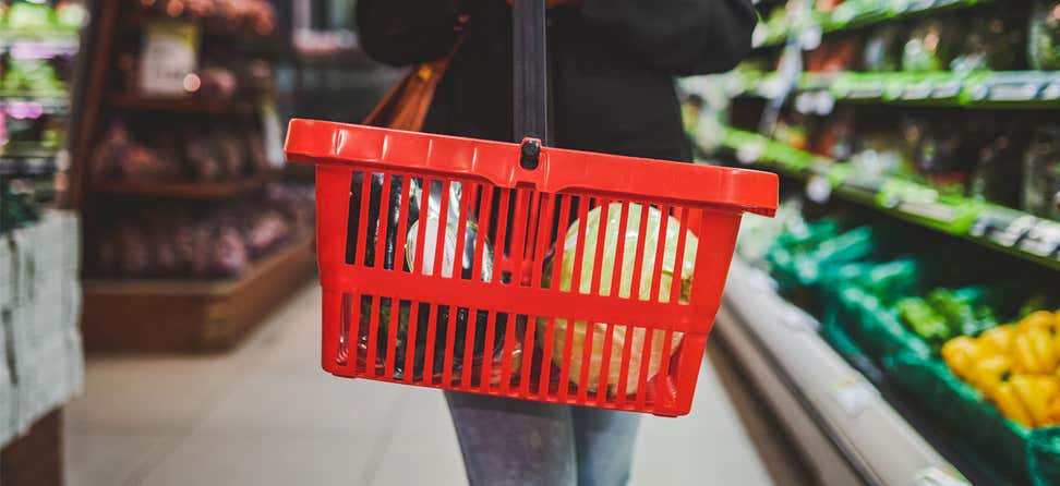 A female is holding a red grocery basket with produce in it in a aisle at the grocery store.