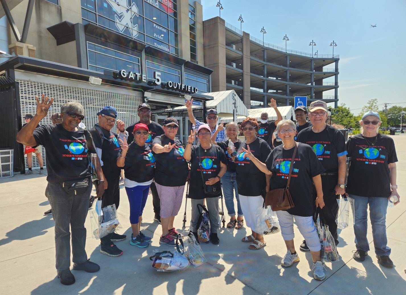 A group of older adults poses outside a stadium, smiling and raising their hands.