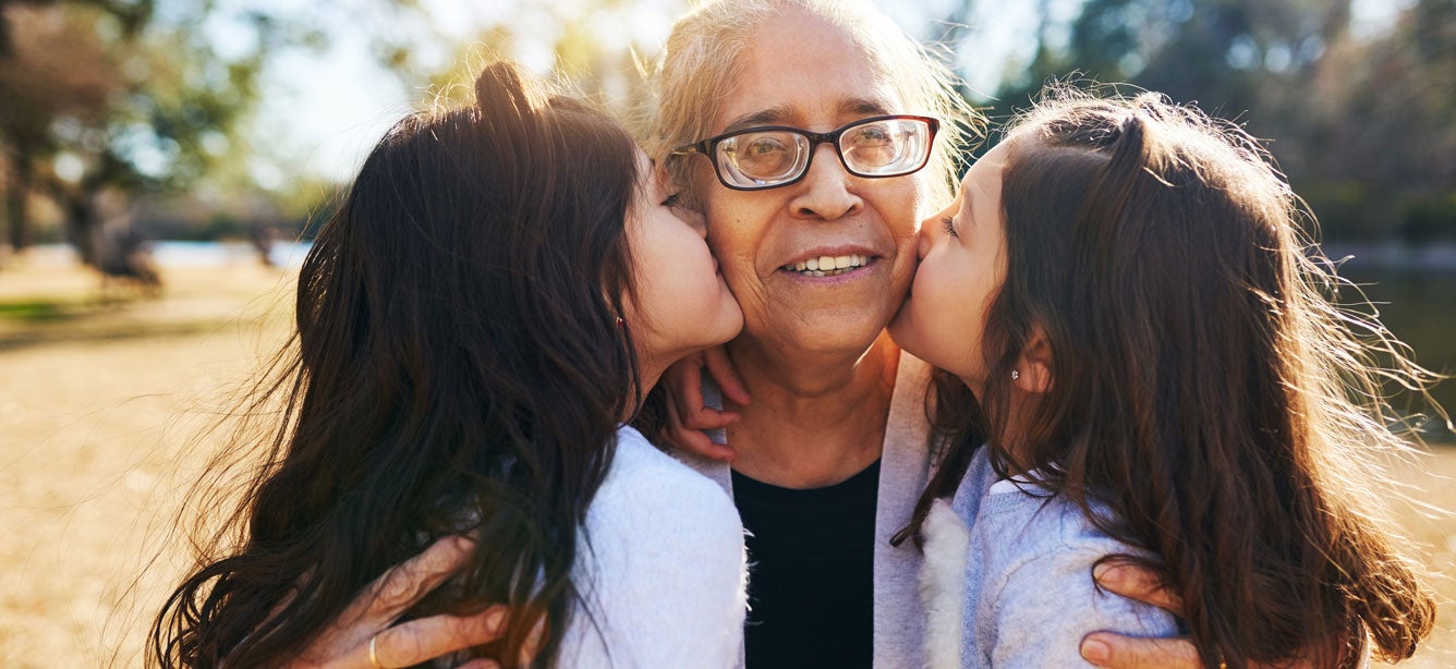 A senior woman is in the parking hugging her granddaughters as they kiss her on the cheeks. 