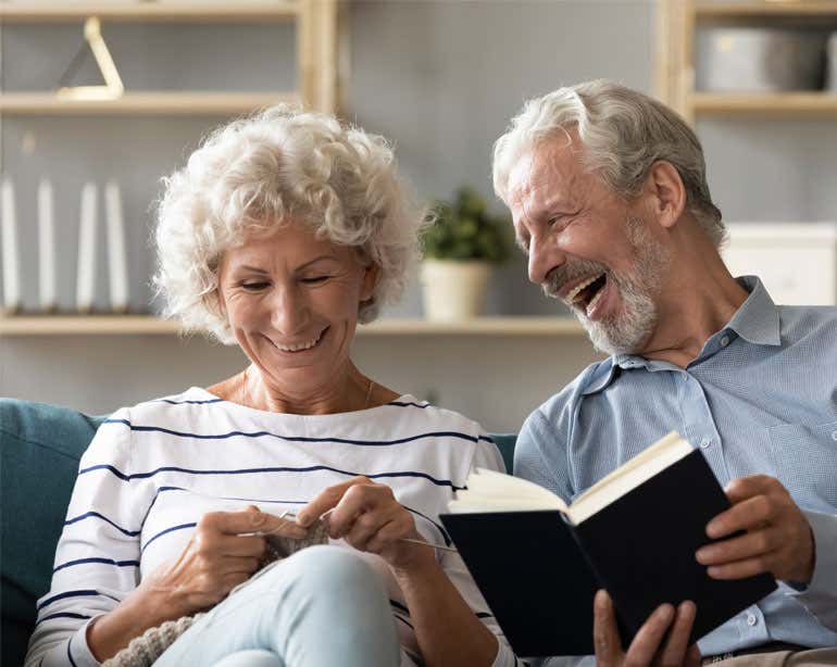 A senior Caucasian couple is sitting on the couch enjoying each other's company - she is knitting, and he's reading a book.