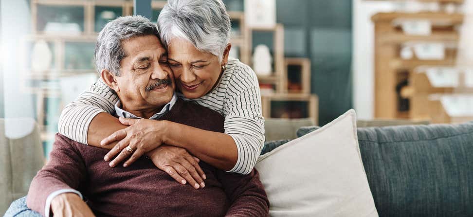 A Black senior woman is hugging her husband from behind while he's sitting on the couch.