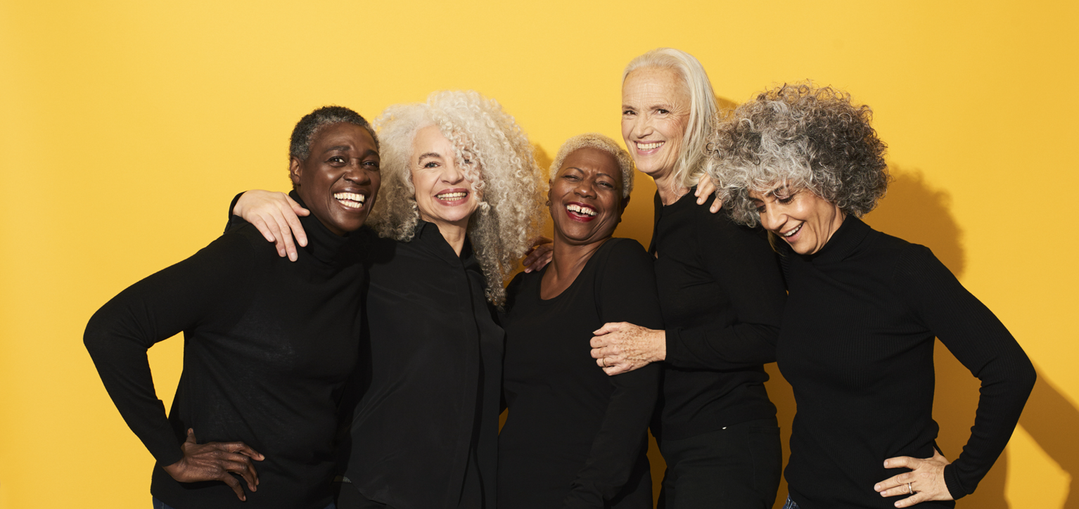 A group of senior women pose for the camera, wearing all black and smiling - with a yellow backdrop.