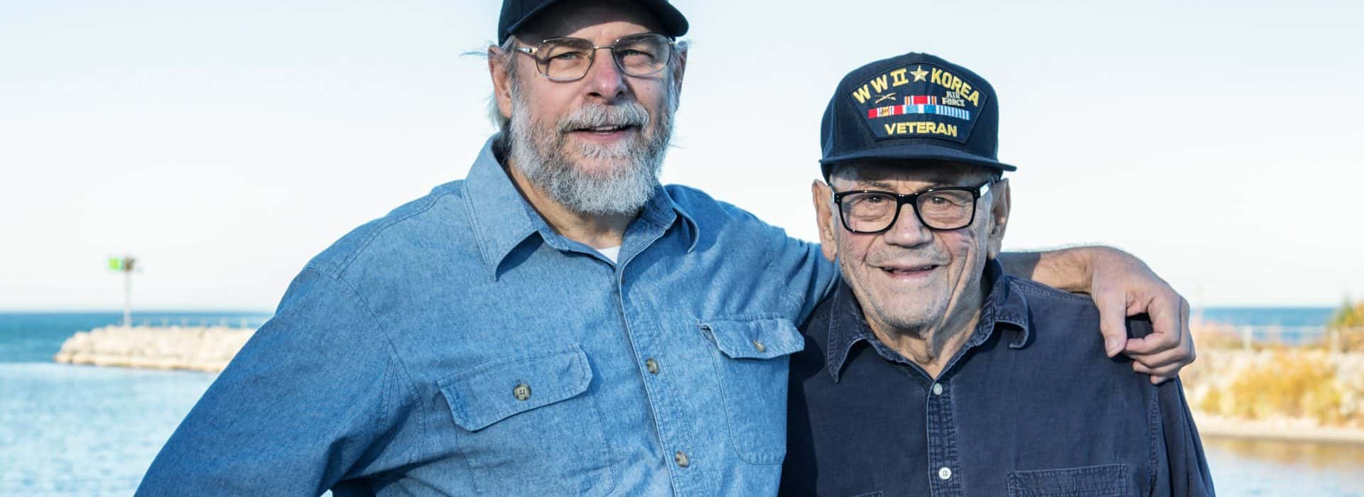 Two elder Vietnam veterans are embracing each other by the shore.