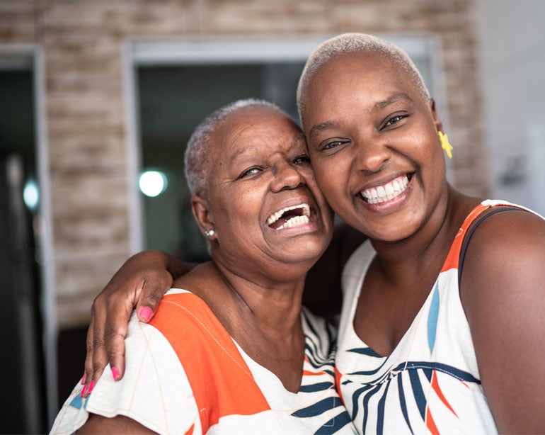A Black senior woman embraces her daughter, both laughing and smiling beautifully.