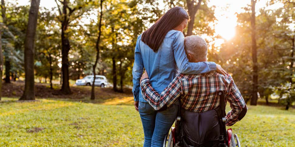 A senior man in a wheelchair is hugging his younger caregiver while they're outside at a park.