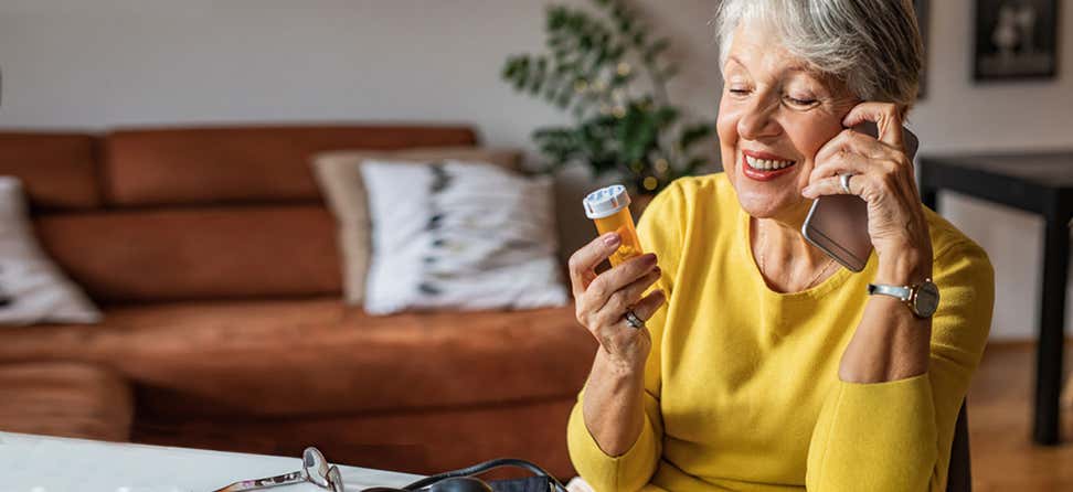 A senior woman wearing a yellow shirt is holding a prescription bottle in her hand while on the phone.