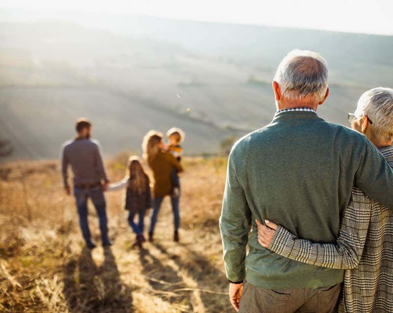 A senior couple hugs at the top of a hill, looking down at their family walking together.